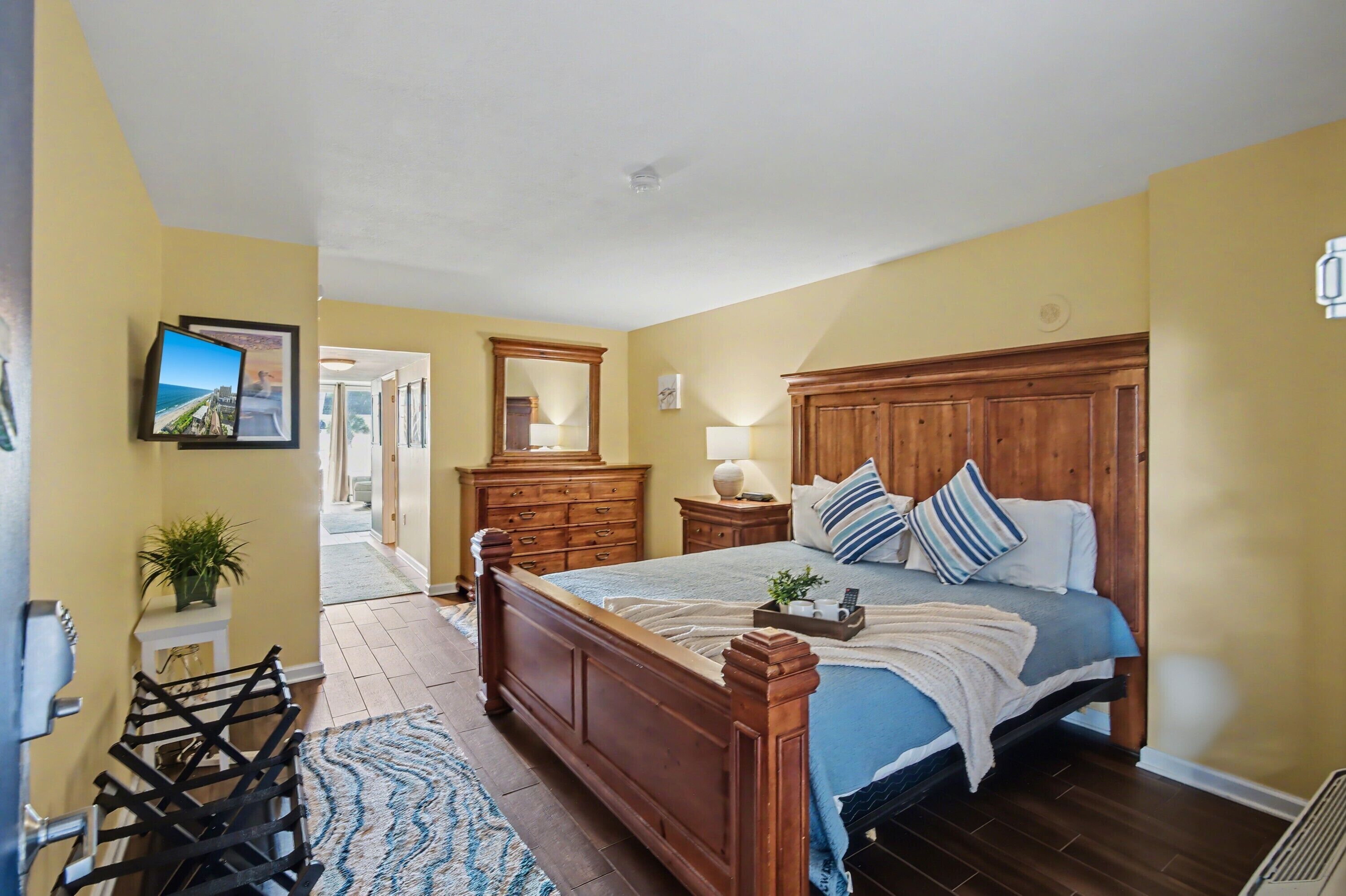 A bedroom with a wooden bed, a nightstand, a dresser, and a mirror. There is a hallway leading to another room.