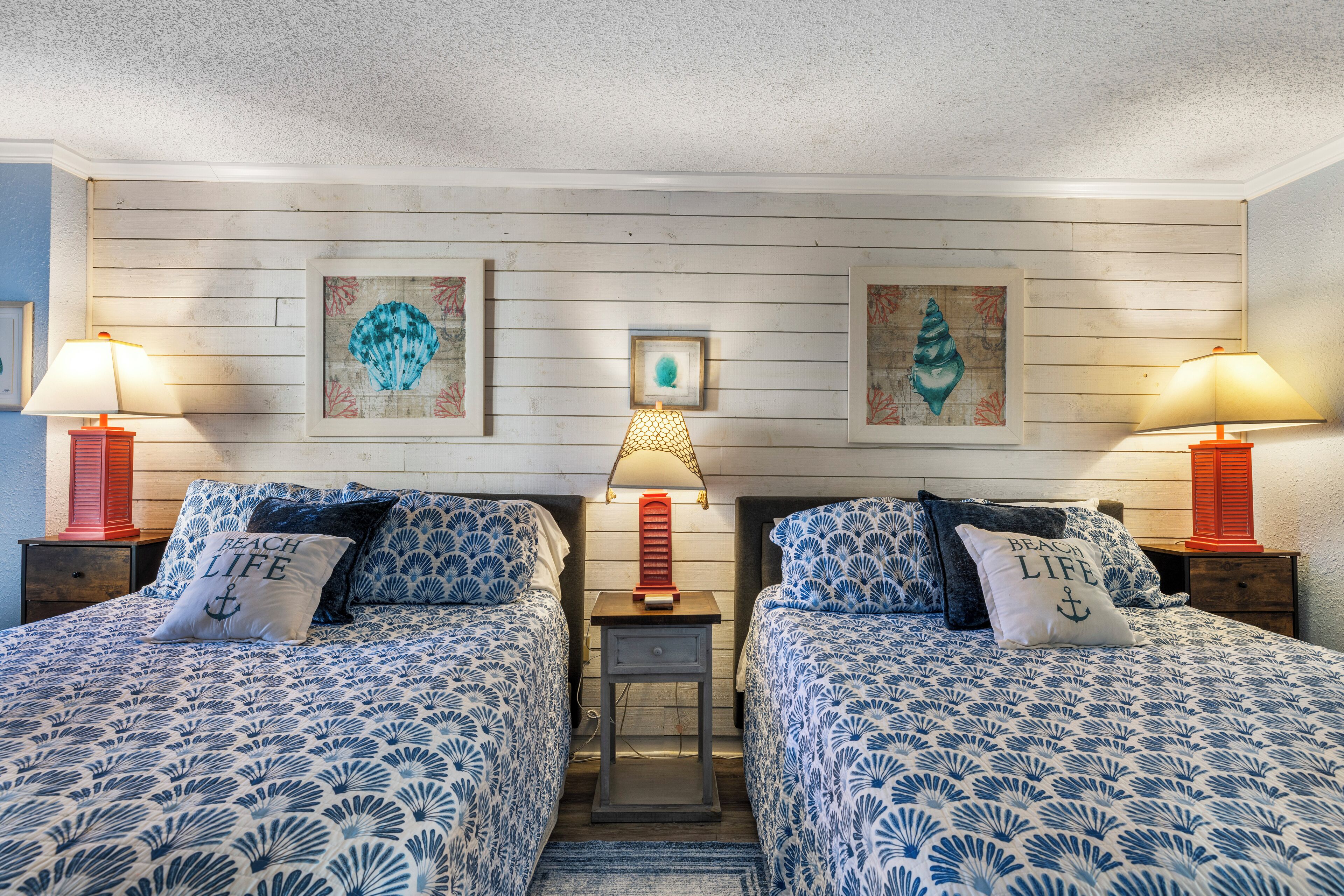 Une chambre avec deux lits, chacun agrémenté de la même literie et des mêmes oreillers, dans des tons de bleu et de blanc. On y trouve deux tables de chevet avec des lampes, et les murs sont décorés d’œuvres d’art encadrées.