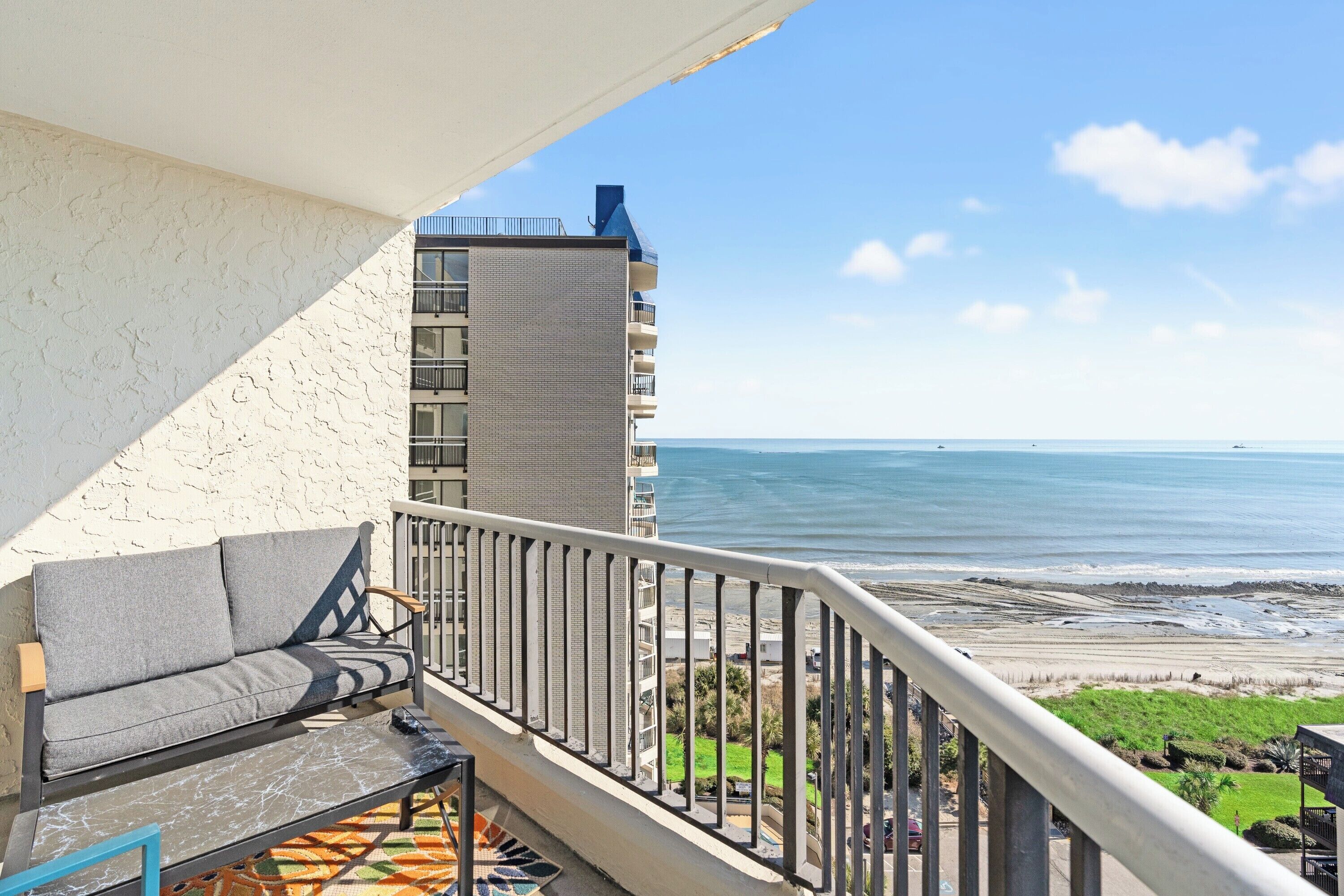 Een balkon met een bank en een tafel, met uitzicht op een strand en een gebouw.