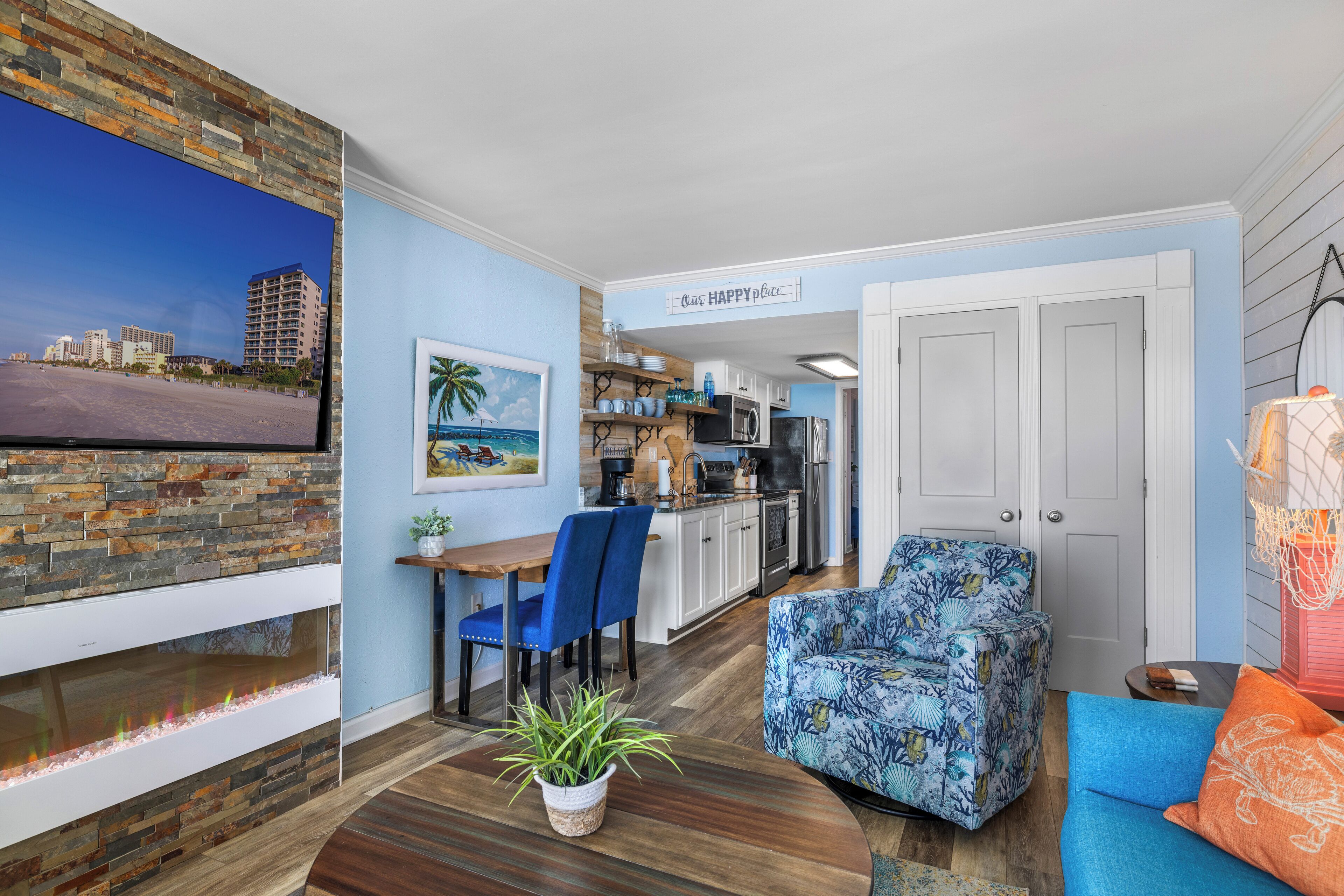 A modern living room with a stone fireplace, a dining area with blue chairs, and a kitchen in the background.