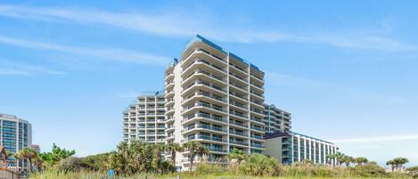 A multi-story hotel with a modern architectural design, located near a beach with visible vegetation and a clear sky.