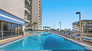 A swimming pool with blue water, surrounded by lounge chairs and a tall building with balconies. - Carolina Winds by Elliott Beach Rentals (Myrtle Beach)