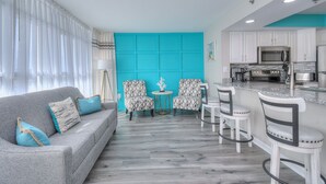 Een moderne woonkamer met een grijze bank, twee fauteuils met een patroon, een kleine ronde tafel, en een keukenhoek met witte kasten en roestvrijstalen apparatuur.