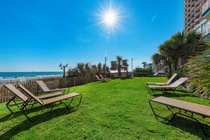 A grassy area with sun loungers facing a beach, a wooden fence, and a building with balconies.