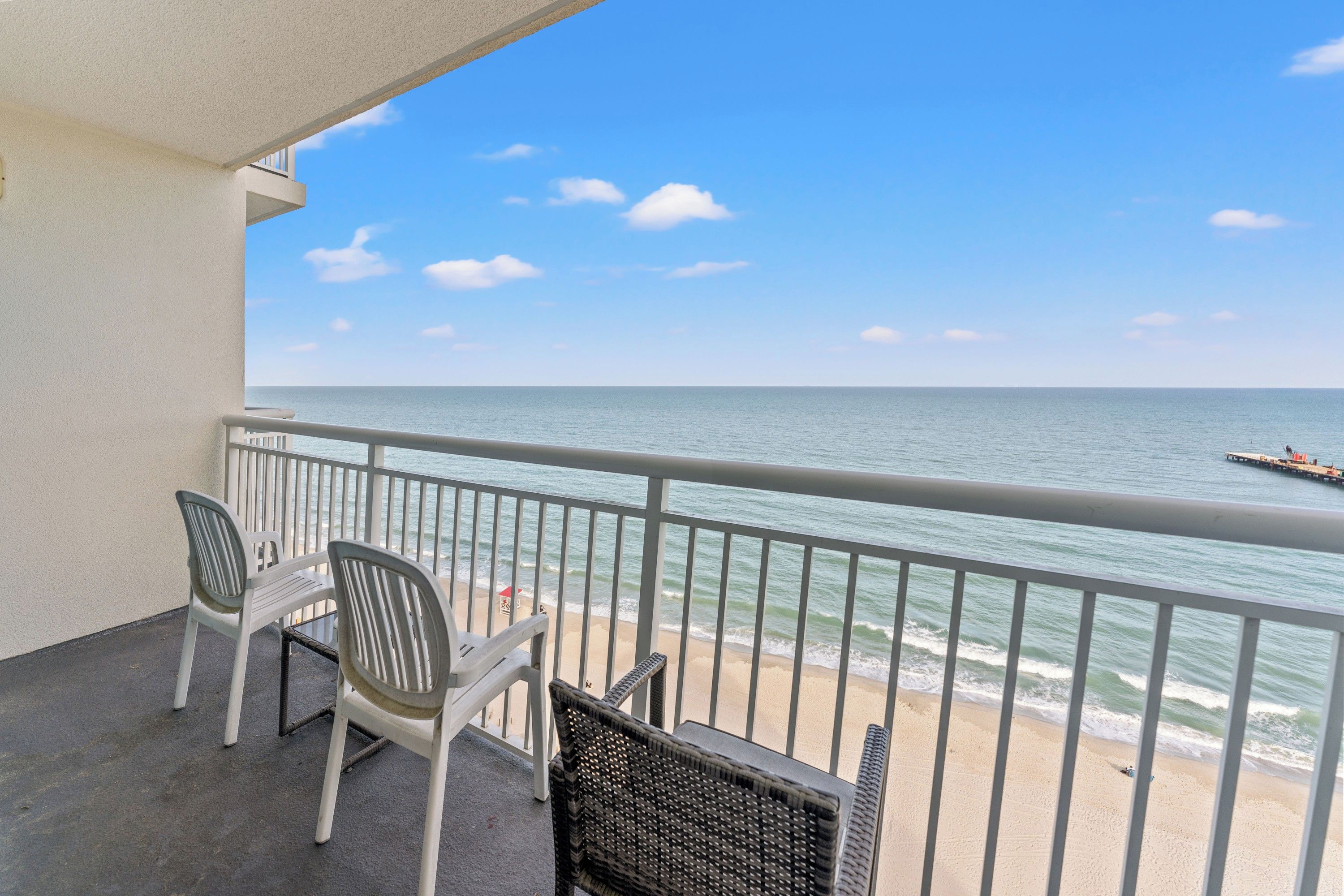 A balcony with two chairs and a wicker chair, overlooking a beach with calm sea and a clear sky.