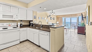 A modern kitchen with white appliances, a granite countertop, and a view to a balcony with seating. - Units at Carolinian Beach Resort by Elliott Beach Rentals (Myrtle Beach)