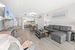 Un salon moderne avec un canapé gris, un coin repas comprenant une table et des chaises, et une cuisine en arrière-plan.