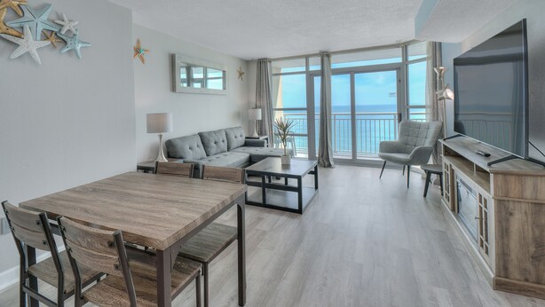 A spacious living room with a large window, a grey sofa, a wooden dining table, and a TV. - Units at Carolinian Beach Resort by Elliott Beach Rentals (Myrtle Beach)