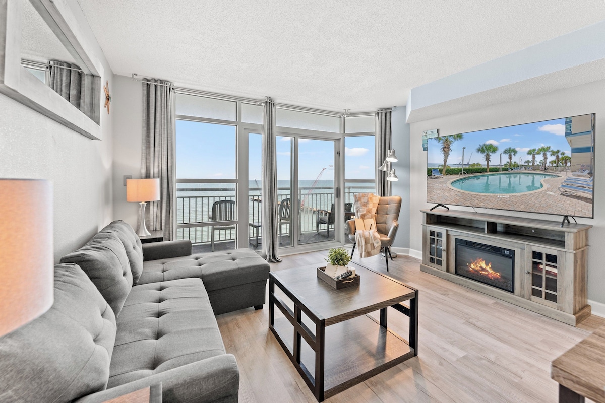 A modern living room with a gray sectional sofa, a coffee table, a fireplace, and a large TV screen displaying a pool view.