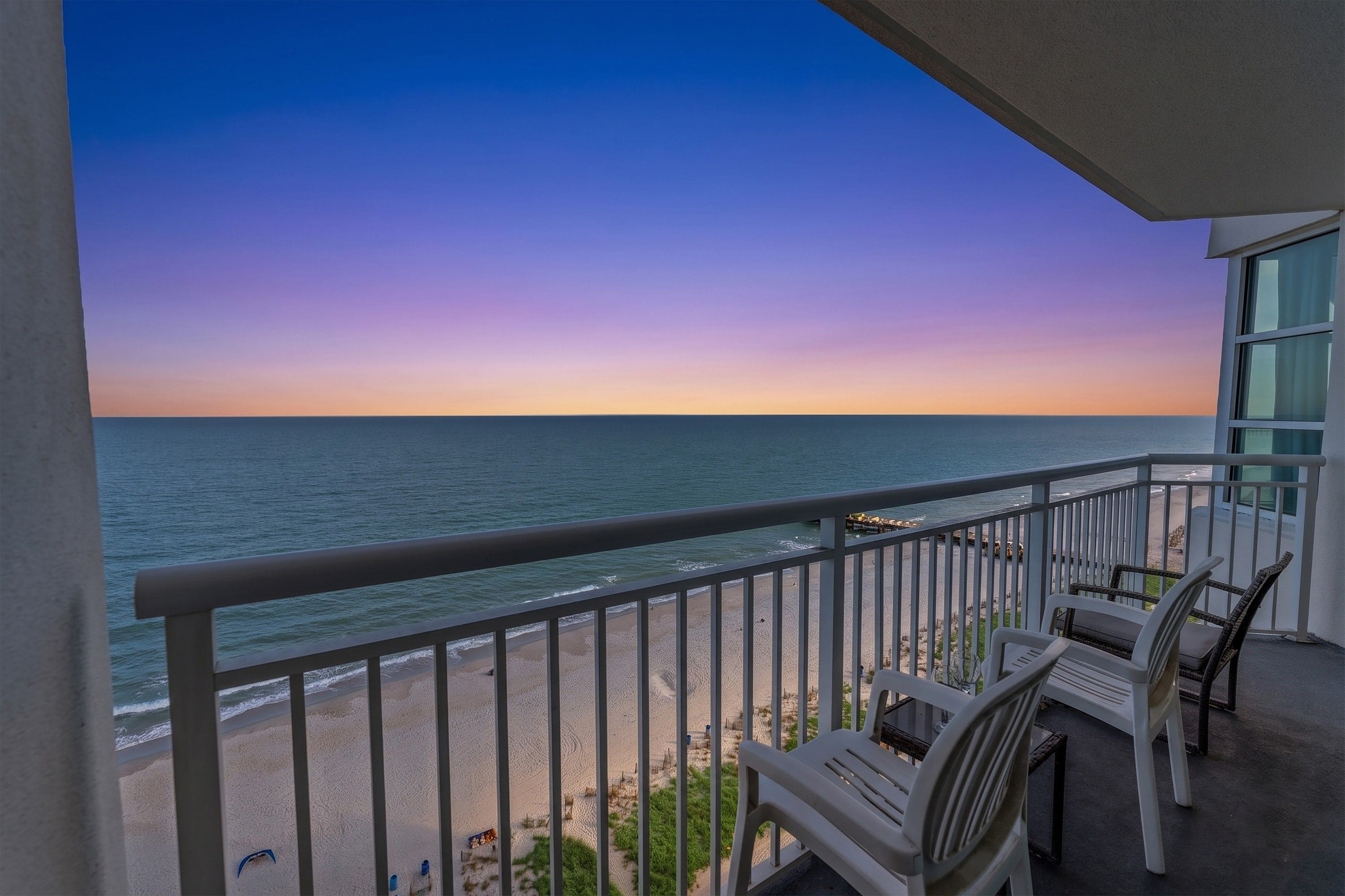 A balcony with a view of the ocean at sunset. There are two white chairs and a table on the balcony.