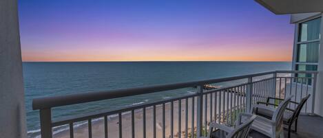 A balcony with a view of the ocean at sunset. There are two white chairs and a table on the balcony.