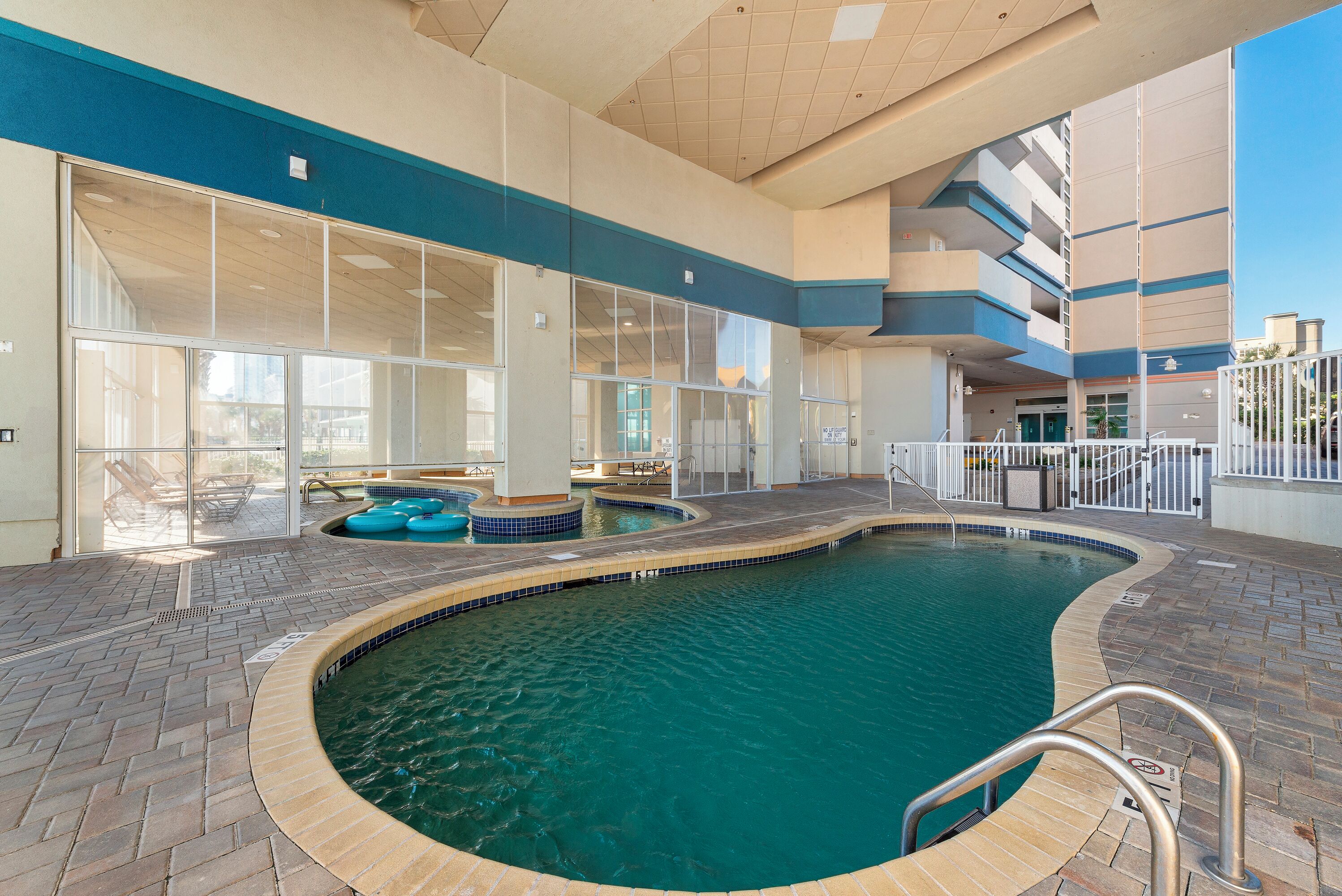 Una piscina interior con paredes de vidrio transparente, un suelo de baldosas y un área junto a la piscina con sillones de descanso.