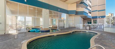 Una piscina interior con paredes de vidrio transparente, un suelo de baldosas y un área junto a la piscina con sillones de descanso.