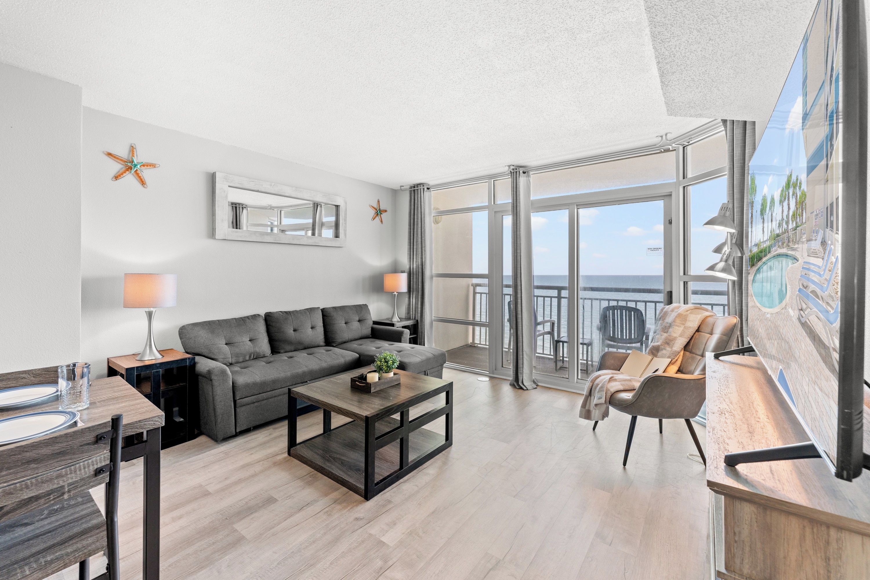 A modern living room with a grey sofa, a coffee table, and a dining area with a wooden table and chairs. There is a balcony with a view of the sea and palm trees.