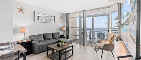 A modern living room with a grey sofa, a coffee table, and a dining area with a wooden table and chairs. There is a balcony with a view of the sea and palm trees.