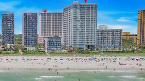 A beachfront hotel with multiple high-rise buildings, a sandy beach with umbrellas, and a clear blue sky. - Units at Carolinian Beach Resort by Elliott Beach Rentals (Myrtle Beach)