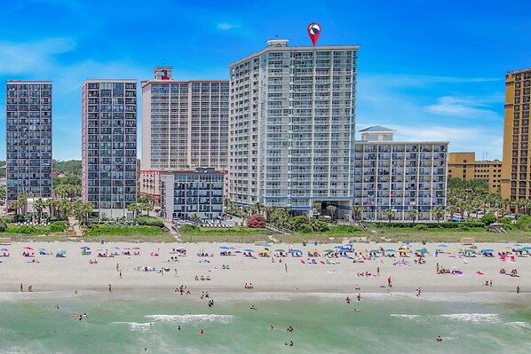 A beachfront hotel with multiple high-rise buildings, a sandy beach with umbrellas, and a clear blue sky.