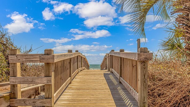 A wooden boardwalk leading to a beach with a clear sky and palm trees.