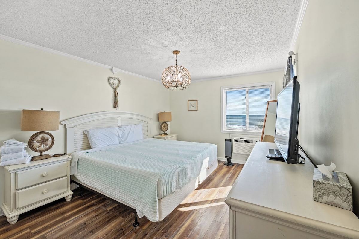 Une chambre à coucher comprenant un lit, des tables de chevet, un bureau, une lampe, un ventilateur, un radiateur et une fenêtre avec des stores.