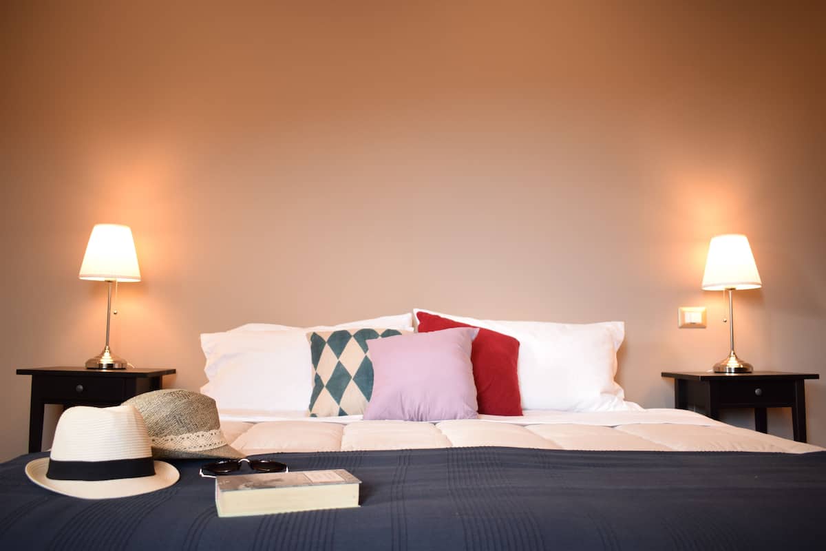 A neatly made bed with white and colored pillows, a hat, and a book on a dark blue bedspread. Two bedside lamps are on either side of the bed.