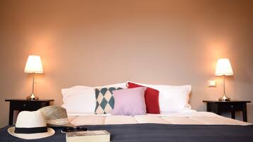 A neatly made bed with white and colored pillows, a hat, and a book on a dark blue bedspread. Two bedside lamps are on either side of the bed.