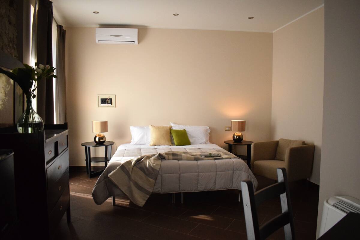 A bedroom with a bed, bedside tables, a chair, a dresser, and an air conditioning unit.