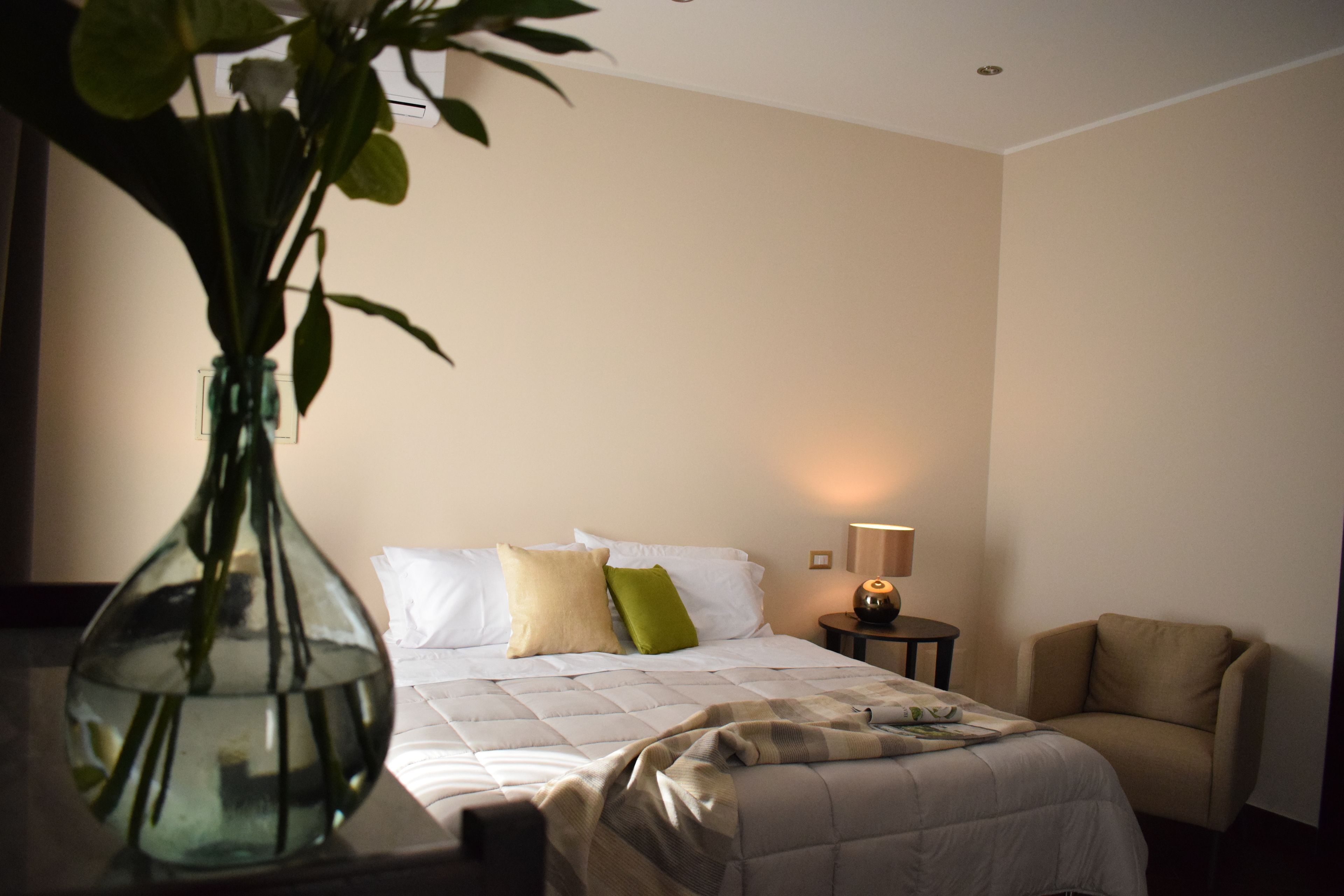 A hotel room with a bed, bedside table, lamp, chair, and a vase with a plant.