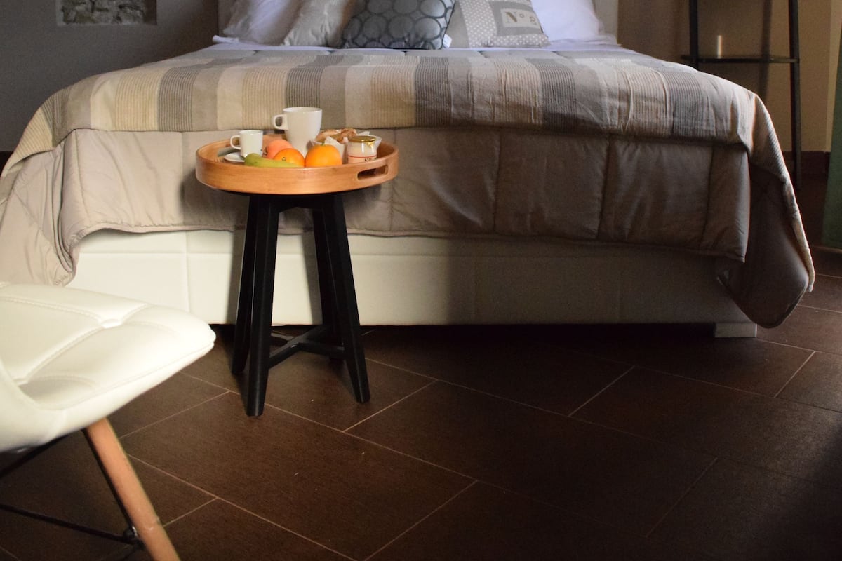 A neatly made bed with a headboard, a bedside table with a tray of fruits and a cup, and a chair.