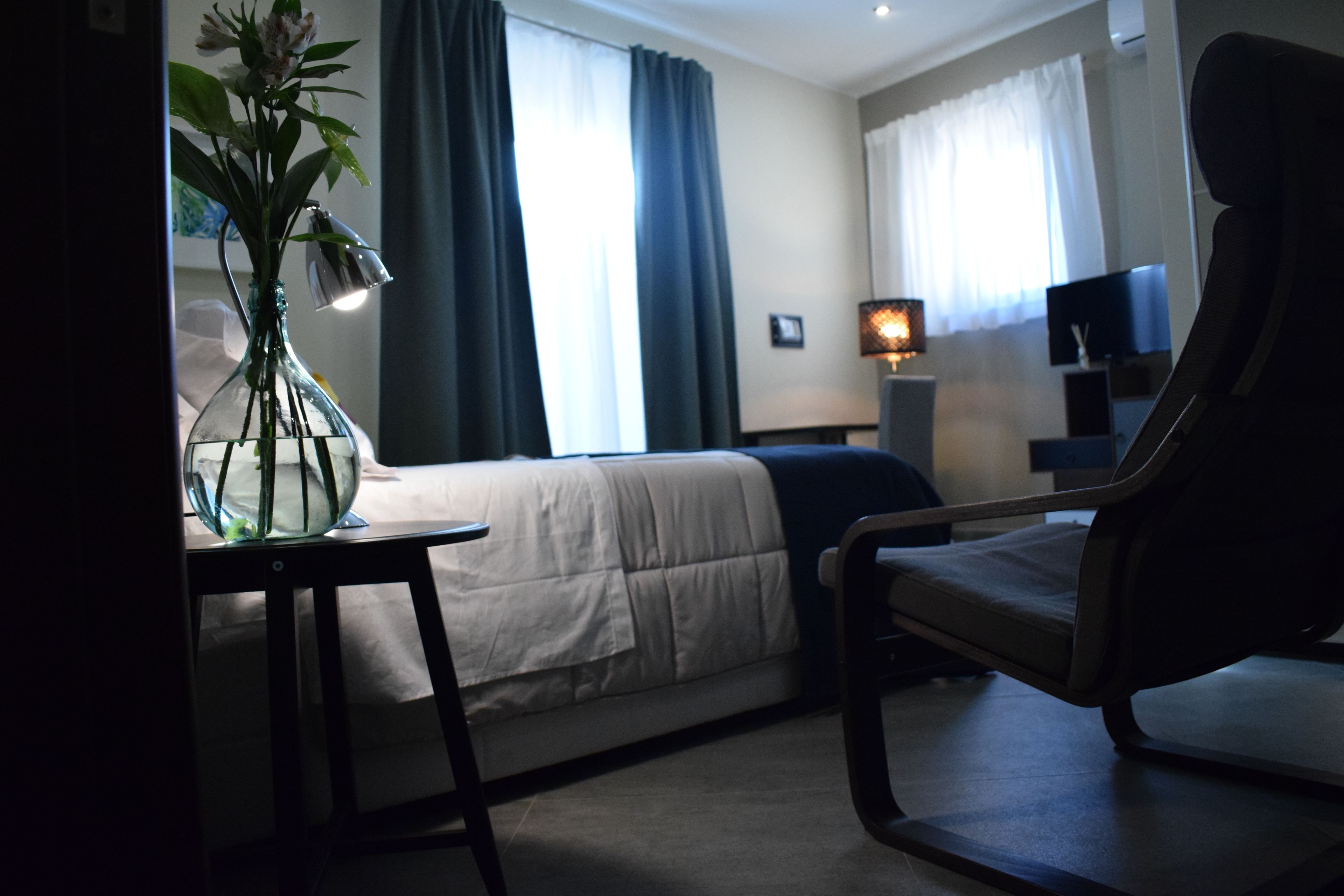 Una habitación de hotel con una cama, una silla, una mesa con un florero y una lámpara.