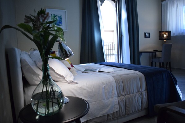 A hotel room with a bed, a bedside table with a lamp, a vase with flowers, and a window with curtains.