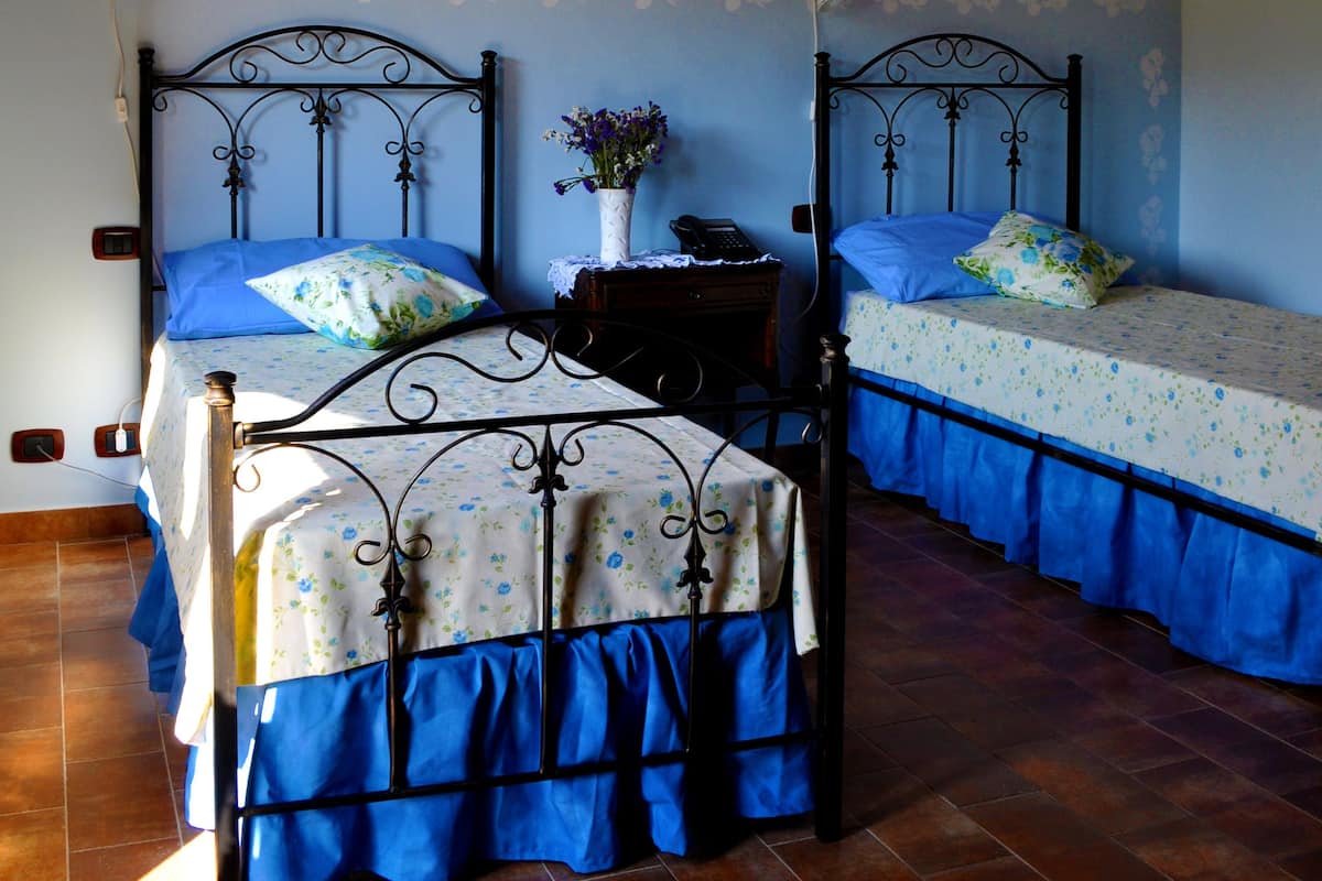 A bedroom with two beds, each with a blue bed skirt and floral bedding. There is a nightstand with a vase of flowers between the beds.