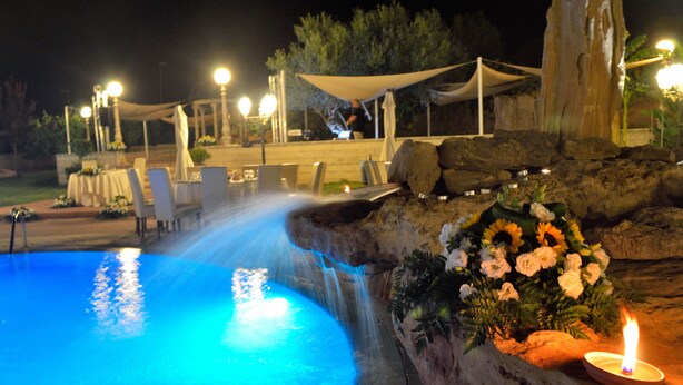 A nighttime outdoor pool area with a water feature, seating, and a decorative flower arrangement. - Tenuta Bellaprima (Niscemi)