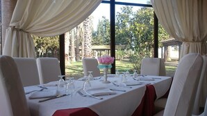 A dining table set for four with white tablecloths and red table runners, surrounded by white chairs. A pink flower centerpiece is on the table. - Tenuta Bellaprima (Niscemi)