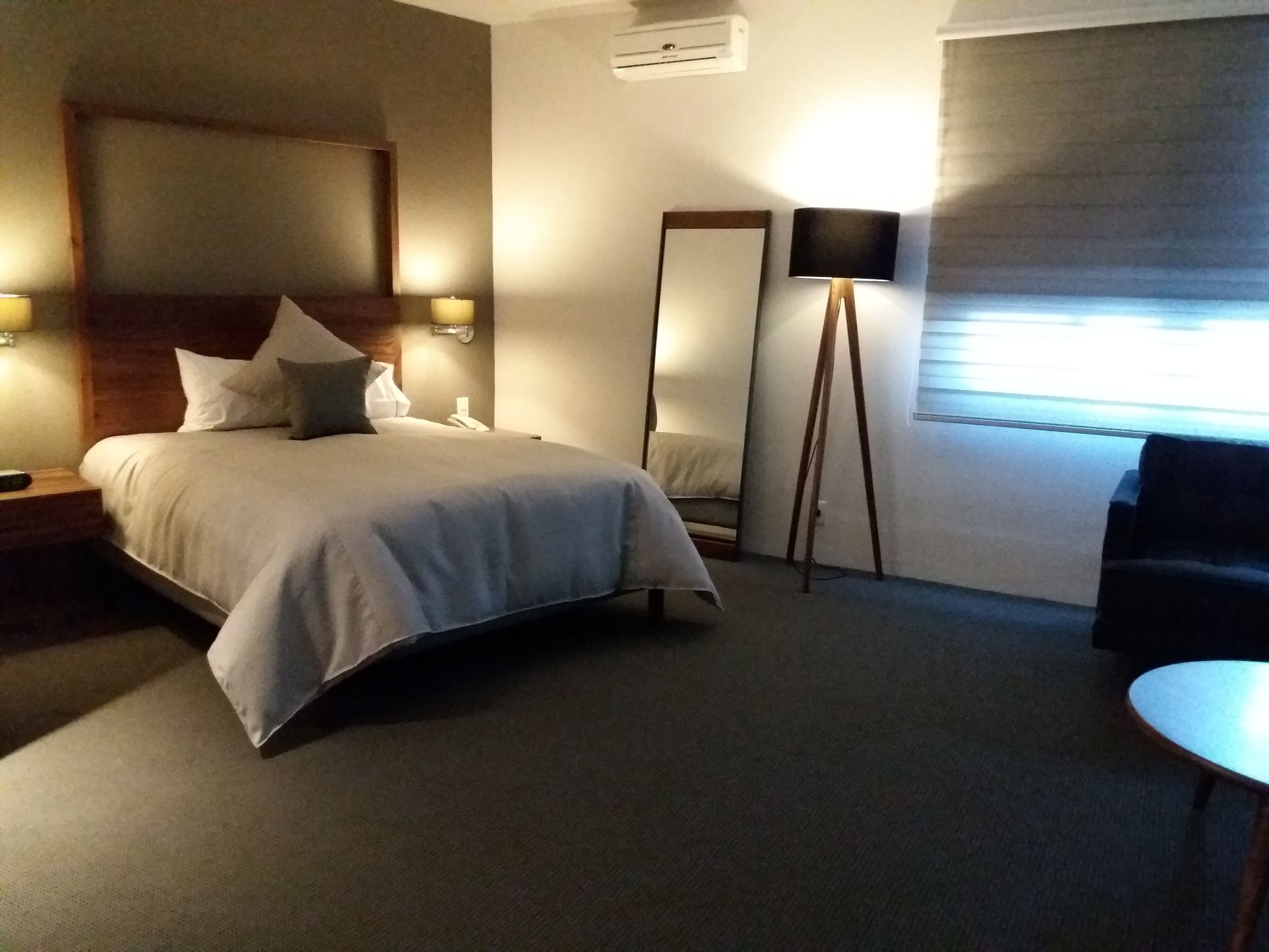 A hotel room with a bed, bedside lamps, a standing lamp, a mirror, a chair, and a small table.