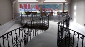 Un escalier en colimaçon orné de rampes en métal ouvragé, menant à un balcon d’où l’on peut admirer un espace extérieur aménagé avec des sièges et de la verdure.