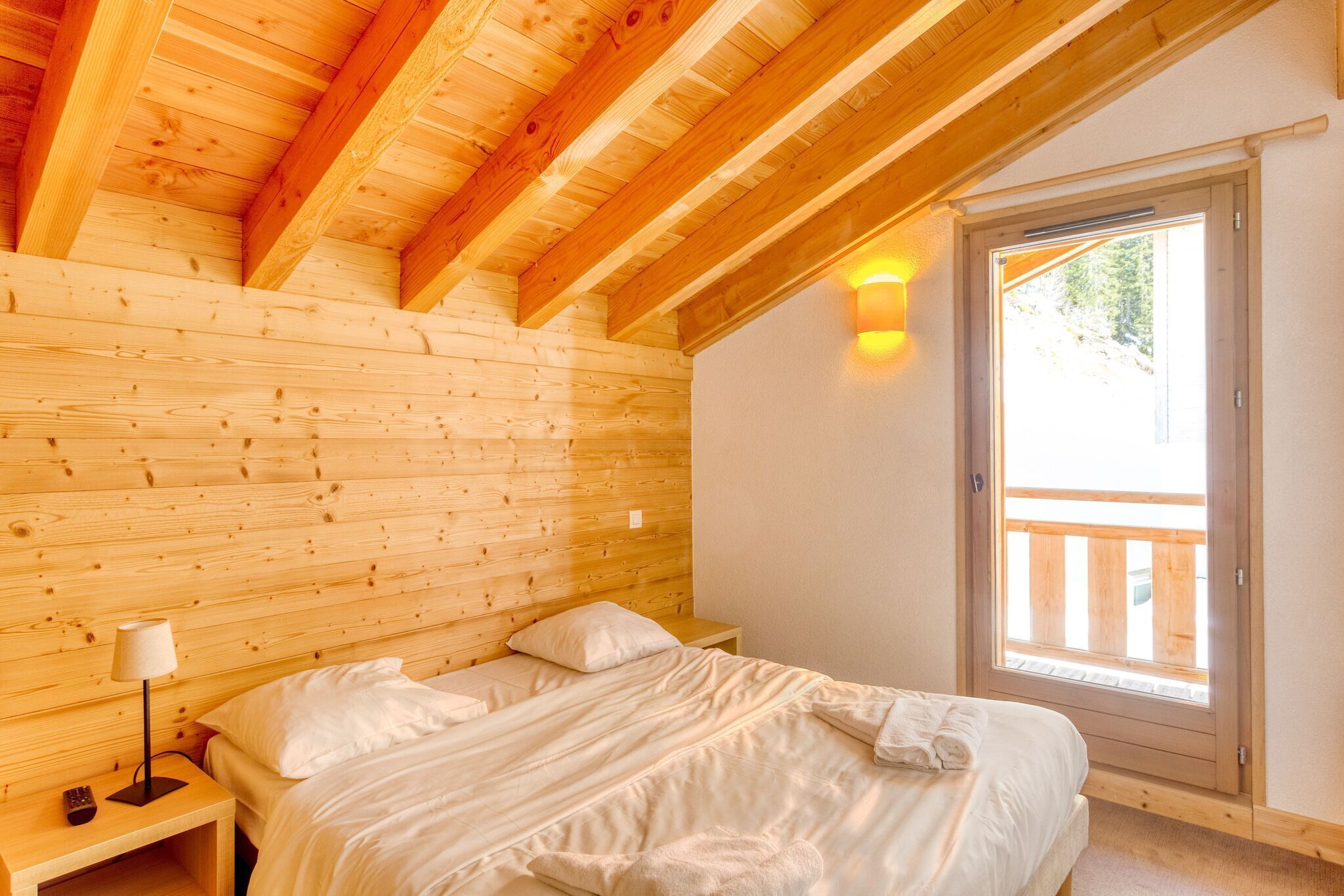 A wooden-paneled bedroom with a bed, nightstand, lamp, and a window with a view of greenery.