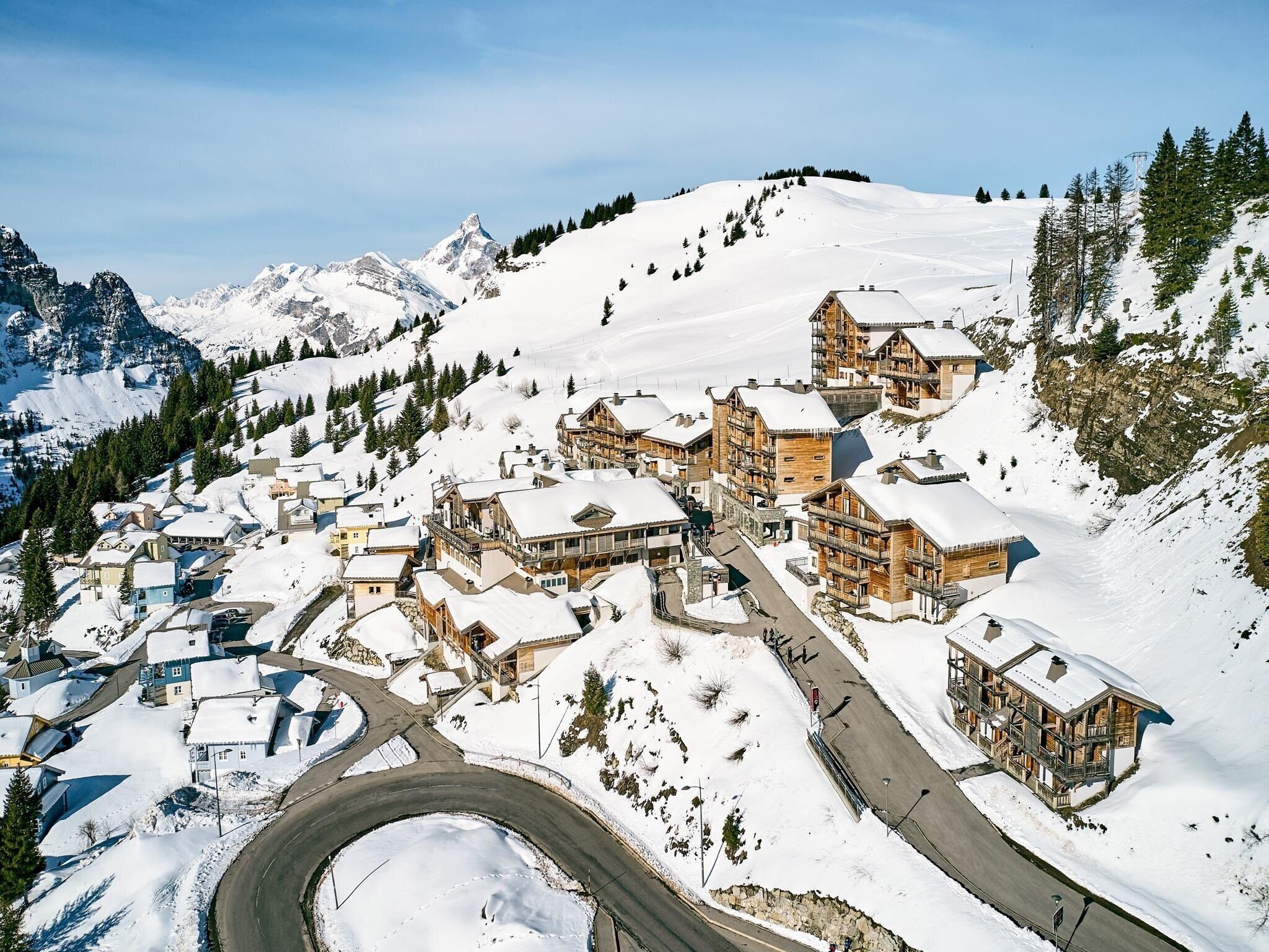 Un village de montagne enneigé, avec des bâtiments en bois, une route sinueuse et une vue dégagée sur les sommets environnants.