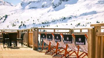 Une terrasse en bois avec des sièges et des bannières Heineken, offrant une vue sur un paysage montagneux enneigé.