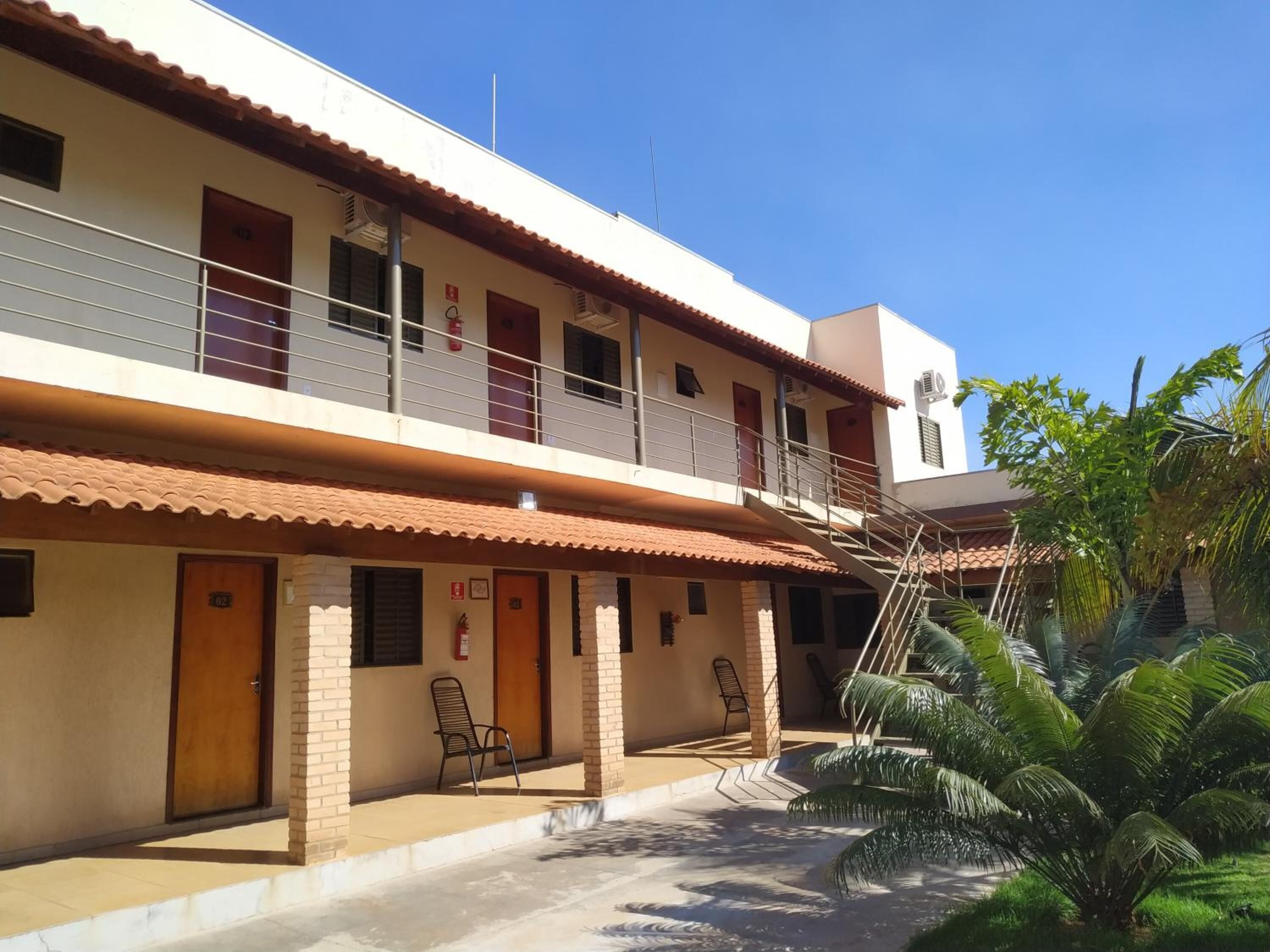 Um edifício de dois andares com uma varanda no andar superior, uma entrada coberta com uma porta identificada com o número '02', e uma pequena área externa com cadeiras.
