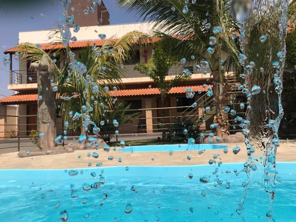 Piscina com água espirrando, cercada por palmeiras e um edifício com telhado de telhas vermelhas.