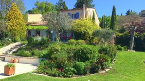 A well-maintained garden with a variety of plants and trees, a stone pathway leading to a building with a tiled roof, and a clear blue sky. - Verdon Views: Pool & Terrace (Montagnac Montpézat)