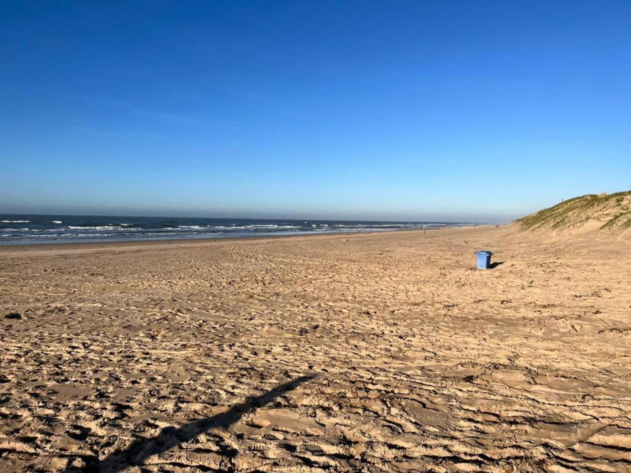 A wide sandy beach with gentle waves, a clear sky, and a dune on the right.