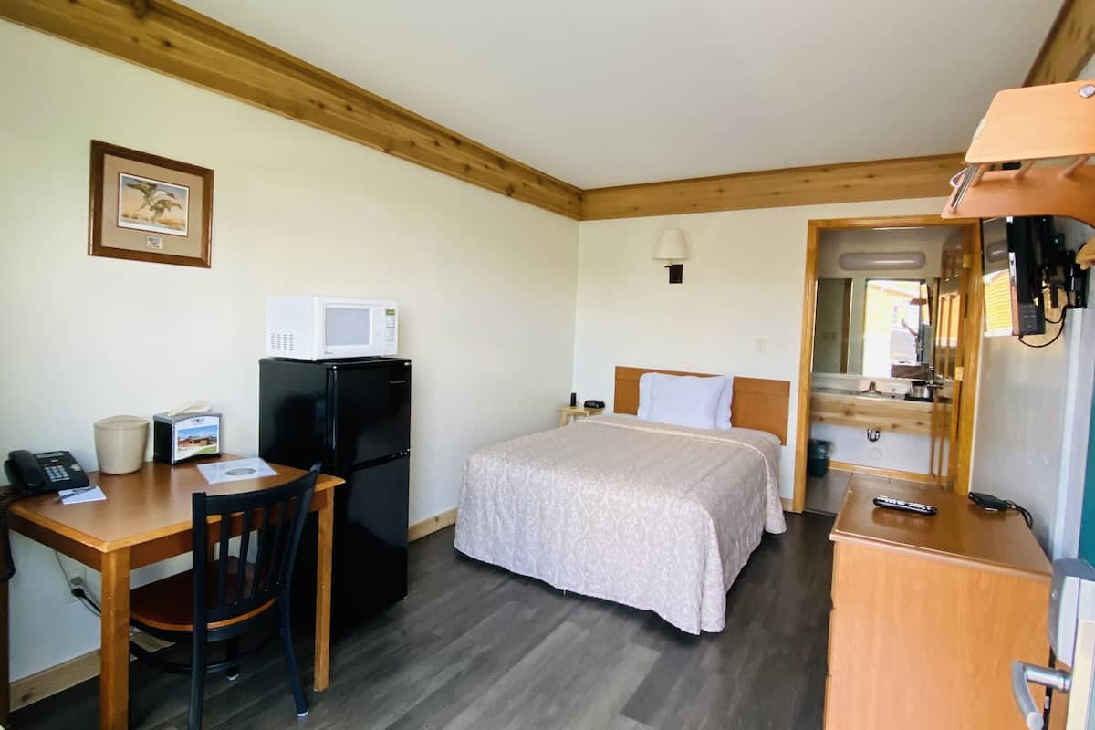 Une chambre d’hôtel compacte avec un plafond en bois, un lit recouvert d’une couverture beige, un bureau avec une chaise, un réfrigérateur, un four à micro-ondes et un petit placard.