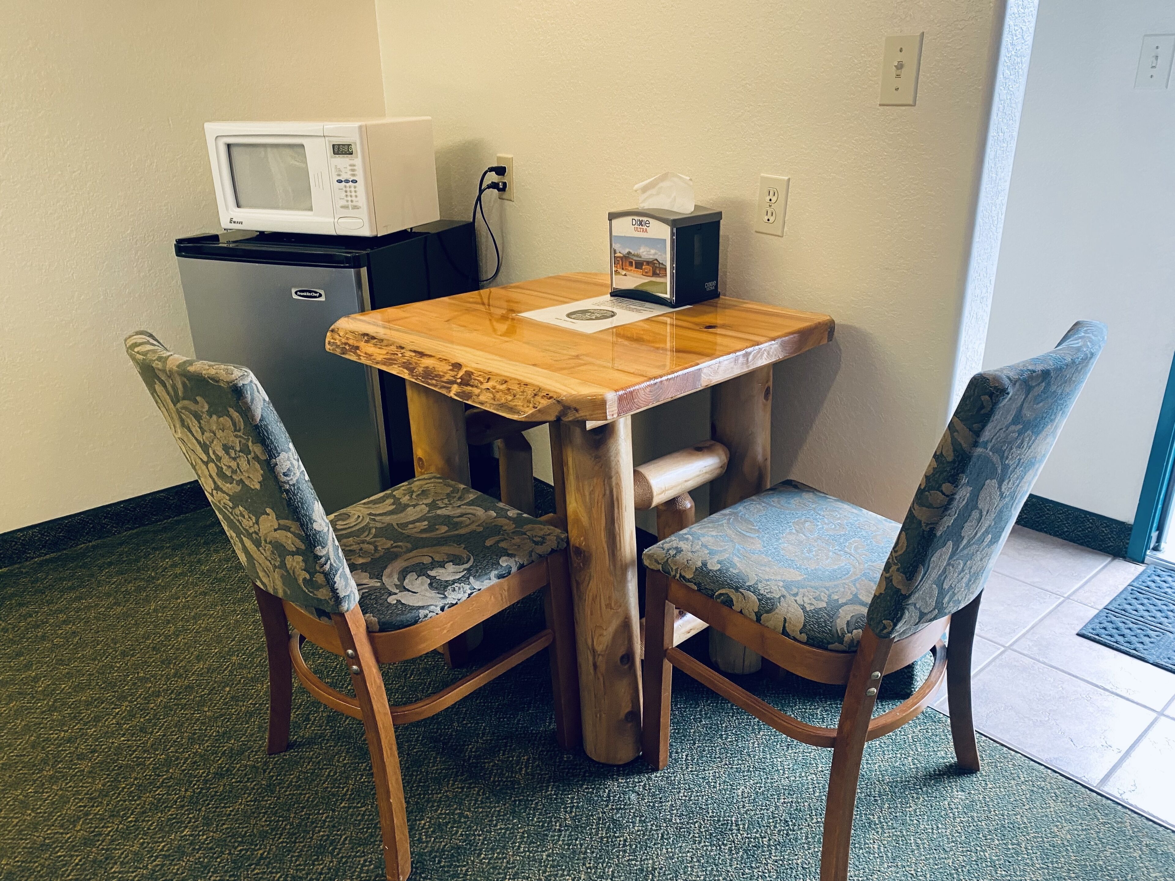 A small wooden table with two upholstered chairs, a microwave, and a tissue box on the table.