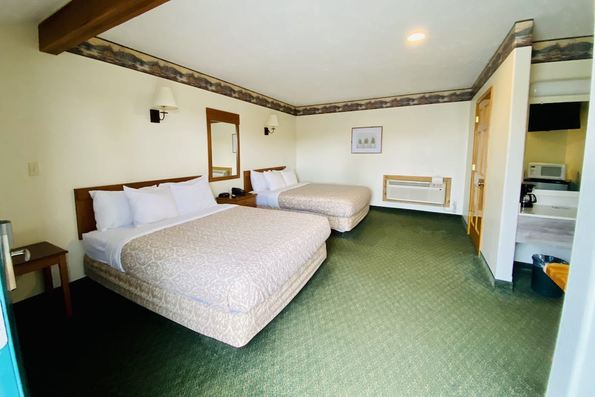 Une chambre d’hôtel avec deux lits, un plafond en bois, une petite table de chevet avec une lampe, un tableau encadré au mur et un coin cuisine.