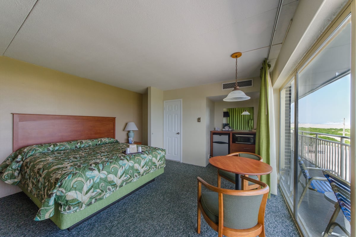 Quarto de hotel com cama, uma mesinha com cadeiras, varanda com vista e cozinha compacta.