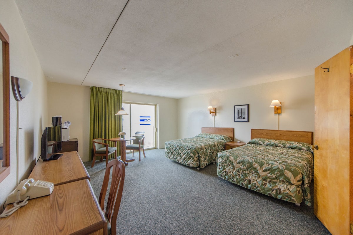 Quarto de hotel com duas camas de solteiro, uma escrivaninha de madeira com telefone, uma cadeira, uma mesinha e uma televisão.