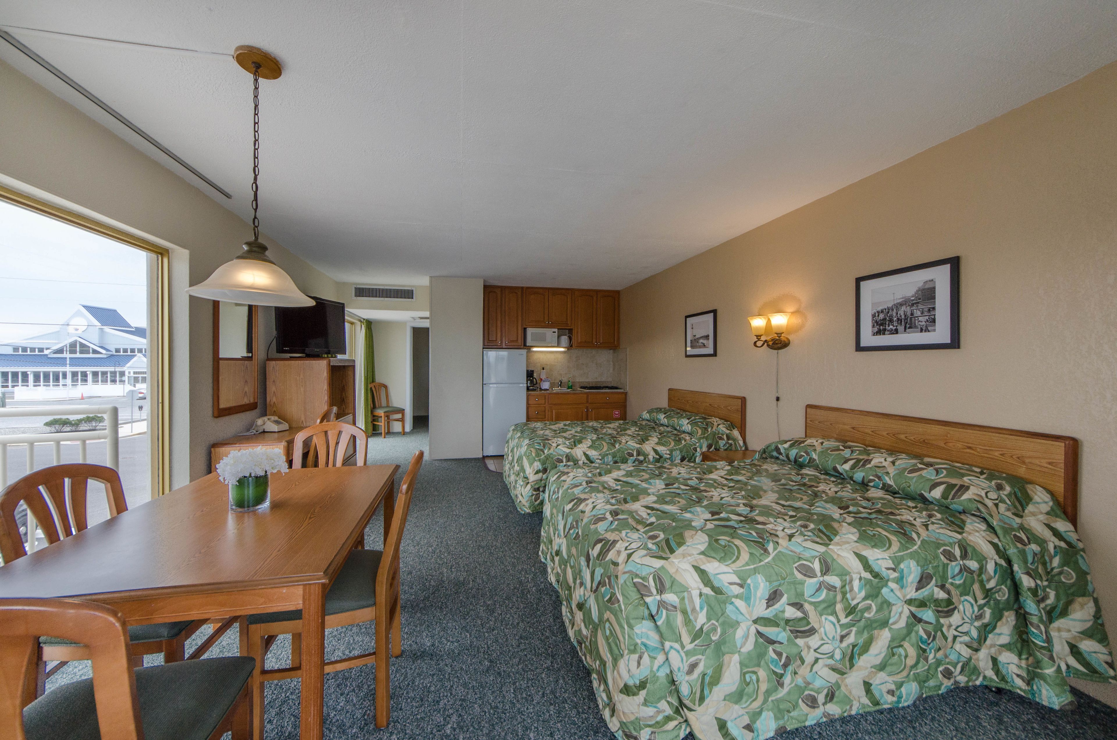 A hotel room with a dining table, chairs, a bed with floral bedding, a kitchenette, and a view of a building outside.
