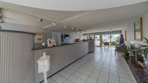 A hotel reception area with a counter, a clock, a telephone, and a seating area with chairs and a plant. - Attache Motel (Wildwood Crest)