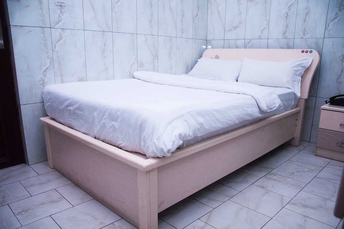 Una cama individual con estructura de madera y ropa de cama blanca en una habitación con paredes de mármol y suelo de baldosas.